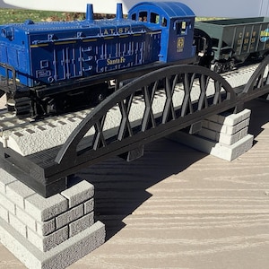May include: A blue Santa Fe model train locomotive and a dark green freight car on a black bridge with a brick-like base. The bridge has a decorative arch design. The image includes the text "A.T.S.F." and "Santa Fe".