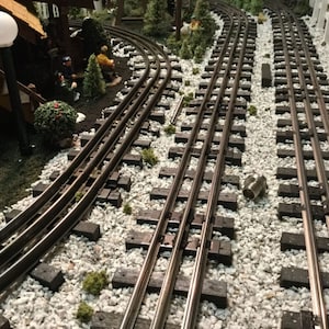 May include: A model train track set with two parallel tracks curving to the right. The tracks are made of metal and are laid on a bed of white gravel. The track is surrounded by miniature trees and a small white picket fence.