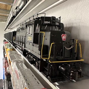 May include: A black model train locomotive with the number 2204 on the front and sides. The train has yellow accents and the Pennsylvania Railroad logo. It is displayed on a shelf with other model trains.