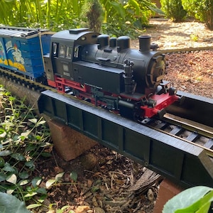 May include: A black toy train engine with a blue boxcar, both on a black bridge over a bed of brown mulch. The engine has a number "80 023" on the side.