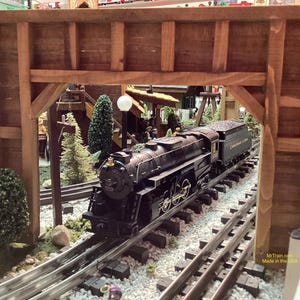 May include: A black model train engine with a gold trim and the words "Chesapeake & Ohio" on the side, traveling on a model train track under a wooden trestle.