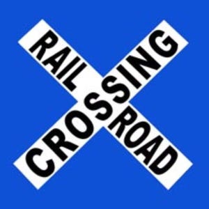 May include: A blue and white railroad crossing sign. The sign is an X shape with the words "RAIL CROSSING ROAD" in black, angled across the sign.
