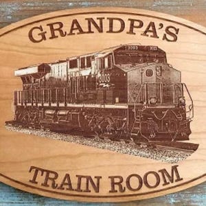 May include: Oval wooden sign with the words "GRANDPA'S TRAIN ROOM" and a detailed engraving of a train engine. The sign is light brown with two pre-drilled holes for hanging.