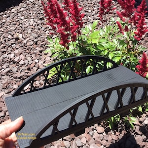 May include: Black metal bridge model with a lattice design. The bridge is arched and has a flat top. The bridge is sitting on a bed of brown gravel.