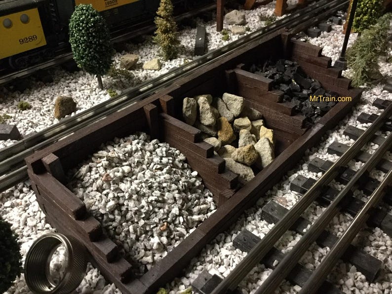 May include: A model railroad scene with a wooden coal hopper filled with black coal and white gravel. The hopper is made of dark brown wood and has a weathered look. The scene is set on a bed of white gravel and includes a section of track.