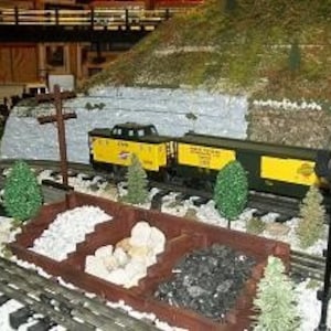 May include: A model train set with a yellow and black locomotive pulling a yellow and black boxcar. The train is on a track with a white gravel bed. There is a small wooden structure with a pile of white gravel and a pile of black coal in the foreground.