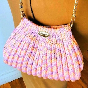 May include: A handmade, knitted handbag in shades of pink, purple, and peach. The bag features a gold-tone chain strap and a small, oval-shaped metal tag with the word "Handmade" on it. The bag's design is ribbed, giving it a textured appearance.