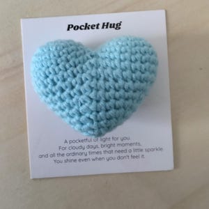 Peut inclure: Un cœur crocheté bleu clair fixé sur une carte blanche avec le texte Pocket Hug en haut et un message imprimé en dessous qui dit A pocketful of light for you. For cloudy days, bright moments, and all the ordinary times that need a little sparkle. You shine even when you don't feel it.