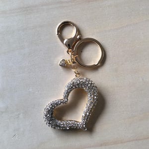 Pode incluir: Um porta-chaves dourado com um pendente em forma de coração cravejado de strass transparentes. O porta-chaves tem um fecho de lagosta e um pequeno berloque de coração. O pendente em forma de coração tem cerca de 5 cm de largura.