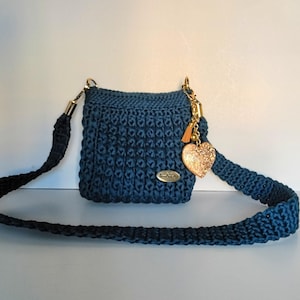 May include: A small, square, navy blue crochet bag with a matching strap. The bag features gold-colored hardware and a charm with a gold heart and tassel. The bag is displayed against a white background.