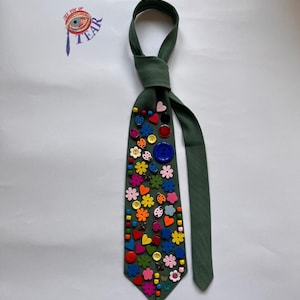 Puede incluir: Una corbata verde oscuro adornada con botones coloridos de varias formas, incluyendo corazones, flores y mariquitas. La corbata tiene un diseño anudado en la parte superior y un extremo puntiagudo. El texto "TEAR" es visible en la esquina superior izquierda.