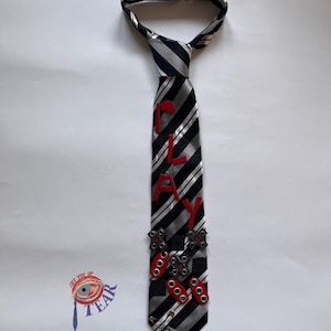 May include: A black and white striped tie with the word "PLAY" in red letters. The tie features red grommets and metal accents. The tie is on a white background.