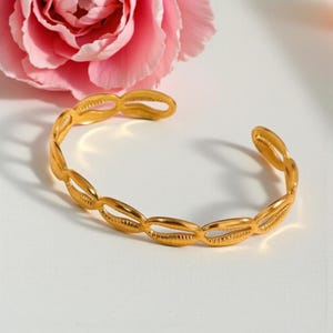 Peut inclure: Un bracelet manchette doré avec un motif de coquillage. Le bracelet ouvert est composé de formes de coquillages liées. Une rose rose est en arrière-plan.