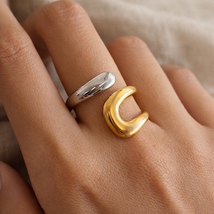 Peut inclure: Deux bagues, une argentée et une dorée, sont portées sur un doigt. La bague argentée a un design lisse et incurvé. La bague dorée a une forme de croissant. Les bagues sont simples et élégantes.