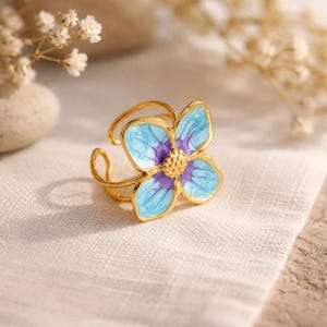 May include: A gold-colored ring featuring a four-petal flower design. The flower has light blue petals with purple accents and a gold center. The ring is set against a white background.