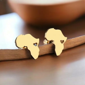 Puede incluir: Un par de pendientes dorados con forma de continente africano, cada uno con un recorte en forma de corazón. Los pendientes se muestran sobre una superficie de madera, con un fondo borroso.