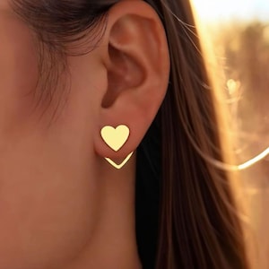 Puede incluir: Primer plano de una oreja adornada con pendientes dorados en forma de corazón. Los pendientes constan de un perno en forma de corazón y un respaldo en forma de V. Los pendientes tienen un acabado dorado brillante, que contrasta con la piel y el cabello.