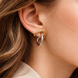May include: A close-up of a gold and silver earring. The upper part of the earring is gold, curving around the ear, while a silver hoop hangs below. The earring is worn on a person's ear, with their skin and hair visible in the background.