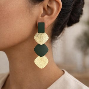 Peut inclure: Une longue boucle d'oreille pendante composée d'un alignement vertical de quatre formes géométriques. Le bijou présente un rectangle vert foncé en haut, suivi d'un losange doré texturé, d'un losange vert foncé lisse et d'un losange doré texturé.