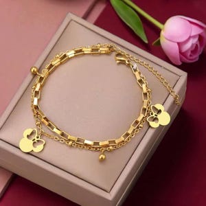 Peut inclure: Un bracelet doré à plusieurs rangs avec un charm en forme de trèfle et de petits accents de boules. Le bracelet est présenté sur une surface neutre avec une fleur rose en arrière-plan. Le bracelet a une fermeture à fermoir.