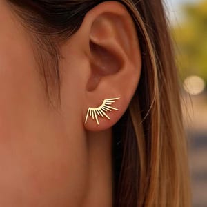 Peut inclure: Boucles d'oreilles dorées en forme d'éclat de soleil. Les boucles d'oreilles présentent un design minimaliste, avec un soleil stylisé et des lignes rayonnantes. Les boucles d'oreilles sont portées sur une oreille, mettant en valeur leur aspect délicat et élégant.