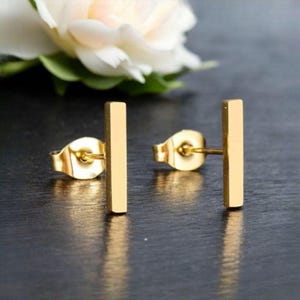 May include: A pair of gold-colored bar stud earrings. The earrings are rectangular and have a simple, minimalist design. The earrings are displayed on a dark surface with a white flower in the background.