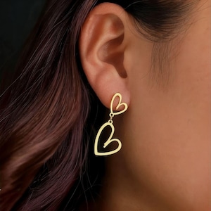 May include: Gold-colored heart-shaped earrings. The earrings feature a small heart connected to a larger heart by a delicate chain. The earrings are worn on a person's ear, with the focus on the jewelry.