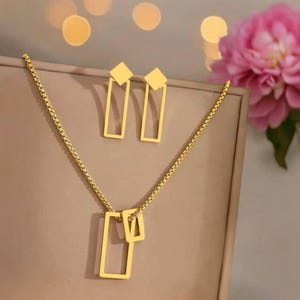 Puede incluir: Un conjunto de joyas de color dorado que incluye un collar y pendientes. El collar tiene una cadena con un colgante rectangular. Los pendientes son rectangulares con una parte superior cuadrada. El conjunto se muestra sobre un fondo neutro.