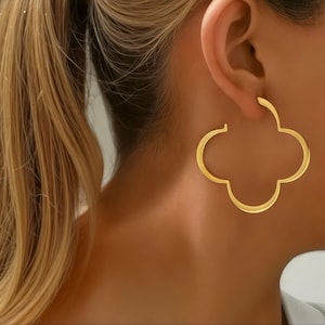 May include: Gold-colored clover-shaped hoop earrings. The earrings are worn on the ear and are a bright, shiny gold color. The earrings have a four-leaf clover design.