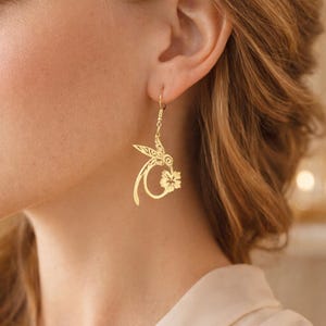 Peut inclure: Boucles d'oreilles dorées en forme de colibri avec un motif floral. Les boucles d'oreilles présentent un design ajouré délicat, avec les ailes et les plumes de la queue du colibri détaillées. Les boucles d'oreilles sont suspendues à un simple crochet.
