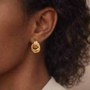 Peut inclure: Gros plan d'une boucle d'oreille dorée. La boucle d'oreille présente un design circulaire avec un nœud torsadé et des barres verticales au-dessus. Le bijou est porté à l'oreille d'une personne, avec le teint visible en arrière-plan.