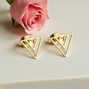Peut inclure: Une paire de boucles d'oreilles dorées avec un motif triangulaire géométrique. Les boucles d'oreilles sont présentées à côté d'une rose rose. Les boucles d'oreilles ont un fermoir à tige.