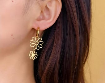 Puede incluir: Pendientes dorados con diseño de margarita. Los pendientes presentan un diseño de aro con dos colgantes de flores de margarita. Las flores están recortadas, revelando la piel que hay detrás.