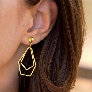 Peut inclure: Boucles d'oreilles pendantes géométriques dorées. Chaque boucle d'oreille présente un petit clou rond en haut, relié à une forme géométrique à double couche et à cadre ouvert. Les boucles d'oreilles sont présentées sur un fond de cheveux bruns et de peau.