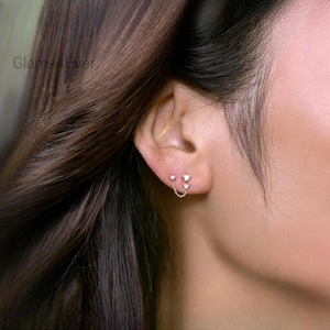 Puede incluir: Pendientes dorados con tres gemas transparentes, unidos por una fina cadena. Los pendientes se muestran en una oreja, mostrando su delicado diseño. Un accesorio elegante.
