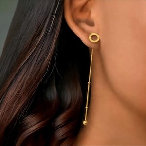 Puede incluir: Pendientes dorados. Los pendientes presentan un pequeño perno circular y una larga cadena colgante con pequeñas cuentas y una bola en el extremo. Los pendientes se llevan en la oreja de una persona, con cabello castaño oscuro en el fondo.