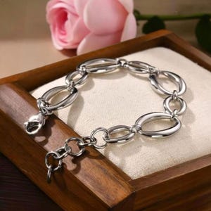 Puede incluir: Una pulsera plateada con eslabones ovalados y un cierre de mosquetón. La pulsera se exhibe en una bandeja de madera con un forro de tela color crema, con una rosa rosa en el fondo.