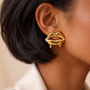Peut inclure: Boucles d'oreilles dorées en forme de lèvres. Les boucles d'oreilles ont un design dégoulinant, ajoutant une touche unique et élégante. Les boucles d'oreilles sont portées sur l'oreille d'une personne aux cheveux foncés et à un blazer blanc.