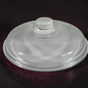 Puede incluir: Una tapa circular de vidrio transparente con un diseño escalonado y una sección central elevada. La tapa incluye un pomo blanco sólido en la parte superior y un patrón de anillo texturizado y esmerilado que rodea el área central.