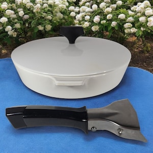 May include: A white enameled cast iron Dutch oven with a black handle on the lid and a separate black and silver handle. The Dutch oven is sitting on a blue surface with a background of white flowers.