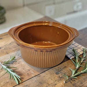 May include: A brown ceramic slow cooker with a ribbed exterior and a smooth interior. The slow cooker has a wide rim with handles and is set on a wooden surface. Rosemary sprigs are placed around the slow cooker.