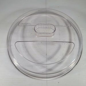 May include: A circular, clear plastic lid featuring a raised, oval-shaped central handle. The surface includes embossed text reading RIVAL on the left side and CLICK-N-LOCK on the right side, with a moulded rim around the outer edge.