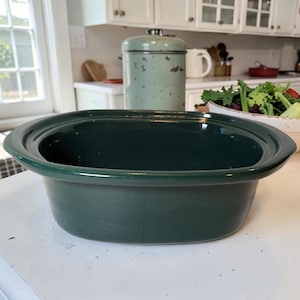 Puede incluir: Una olla de cocción lenta ovalada de color verde oscuro, perfecta para preparar comidas. La olla de cerámica tiene un acabado liso y brillante. El electrodoméstico de cocina está sobre una superficie blanca, con un recipiente verde claro y un cuenco de ensalada en el fondo.