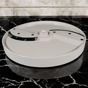May include: A white food processor slicing disc with a circular design. The disc has a central hub and a curved blade. Small black rivets are visible on the disc's surface. The disc is designed for food preparation.