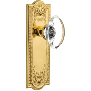 May include: A polished brass door handle with a clear, oval-shaped crystal knob. The rectangular backplate features ornate floral and scrollwork detailing. The handle is designed for interior doors and adds a touch of elegance.