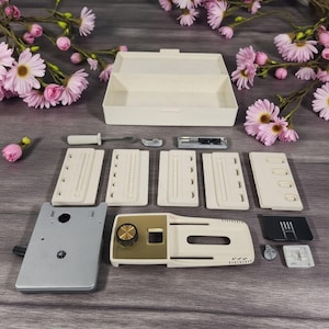 May include: A sewing machine accessory kit with various parts, including a beige storage box, several beige plates, a silver screwdriver, and a silver and gold-toned machine component. The items are arranged on a wooden surface, with pink flowers in the background.