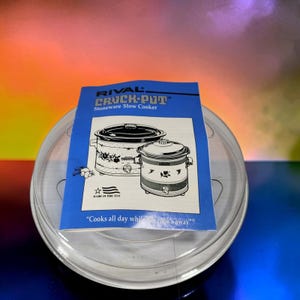May include: A clear, circular glass lid for a slow cooker, topped with a blue and white instruction manual for a Rival Crock-Pot Stoneware Slow Cooker. The manual features illustrations of two slow cookers and the text, "Cooks all day while the cook's away."
