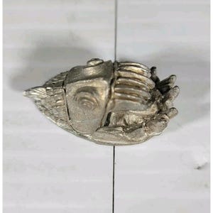 May include: A silver-colored metal sculpture of a fish. The fish has detailed fins, scales, and a segmented body. The sculpture is on a white surface.