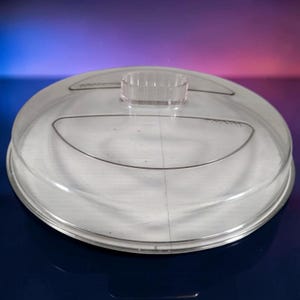 May include: A circular, clear plastic lid featuring a central, raised, oval-shaped handle. The lid has a smooth, transparent surface with a subtle, molded ridge design across the top and the word Rival embossed near the edge of the handle.