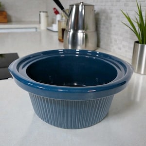 Może przedstawiać: Głęboko niebieski ceramiczny wolnowar z żebrowanym zewnętrzem. Wolnowar ma szeroki, zaokrąglony brzeg i gładkie wnętrze. Stoi na białym blacie.